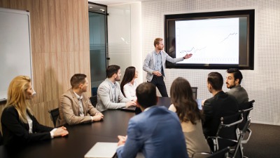 Controller zeigt acht Kollegen im Konferenzraum eine Powerpoint Präsentation mit Entwicklungen der Unternehmenszahlen